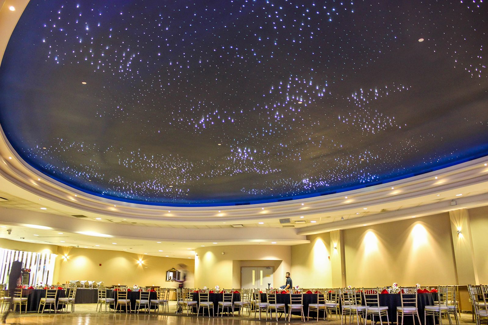 The perimeter of a large banquet room is filled with nearly a dozen round tables draped in cloth and surrounded by chairs. Two thirds of the photo is taken up by the ceiling, which resembles clusters of stars in the night sky. At the back of the room near a door, a person sets one of the banquet tables.