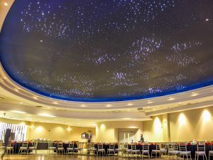 The perimeter of a large banquet room is filled with nearly a dozen round tables draped in cloth and surrounded by chairs. Two thirds of the photo is taken up by the ceiling, which resembles clusters of stars in the night sky. At the back of the room near a door, a person sets one of the banquet tables.