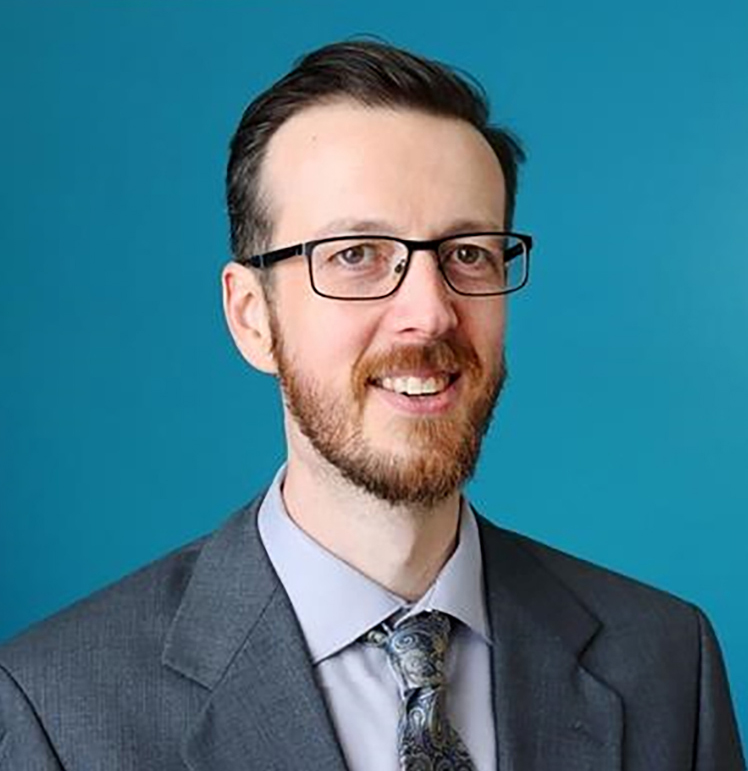 A man with glasses poses for a professional headshot in front of a blue background wearing a grey suit and patterned tie.