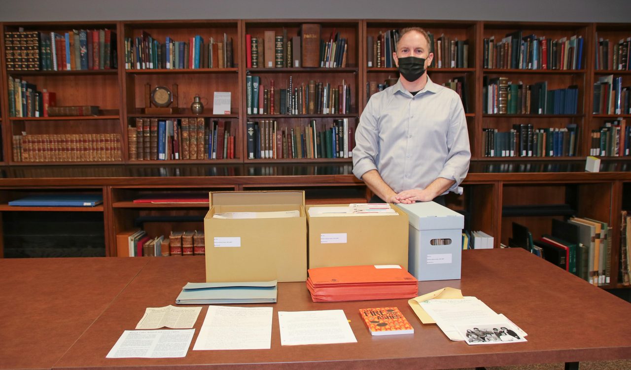 A masked man stands in front of a table filled with boxes and documents, with a row of book shelves in the background.