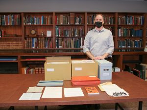 A masked man stands in front of a table filled with boxes and documents, with a row of book shelves in the background.