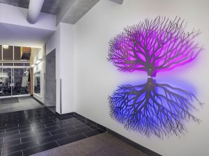 A stainless steel sculpture of a tree and its reflection, back-lit and illuminated purple on top and blue on the bottom, sits mounted on a white wall in an office entry way with concrete ceilings leading towards glass doors leading into the building.