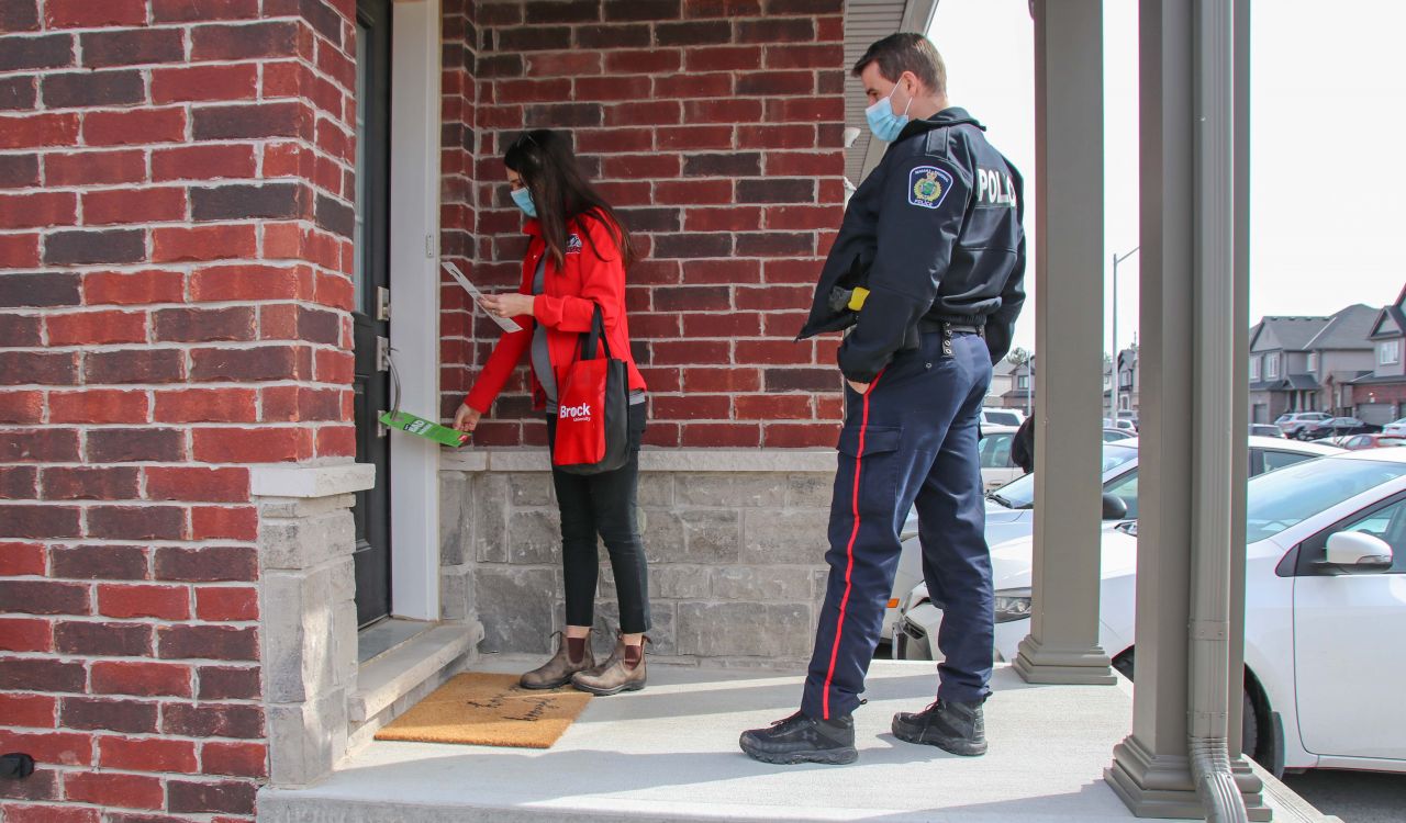 A woman in a red coat hangs a pamphlet on a door handle while she is accompanied by a male police officer.