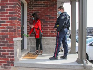 A woman in a red coat hangs a pamphlet on a door handle while she is accompanied by a male police officer.