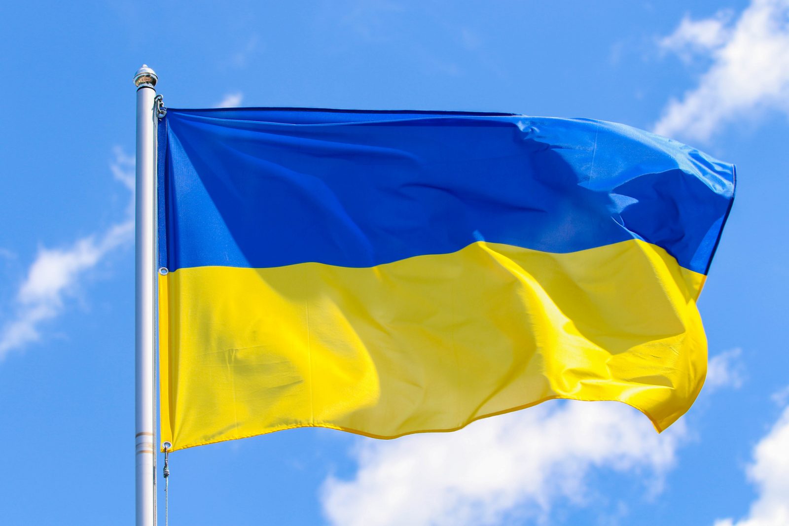 The Ukrainian flag, with blue on the top and yellow on the bottom, flies on the right side of a flagpole against a blue sky.