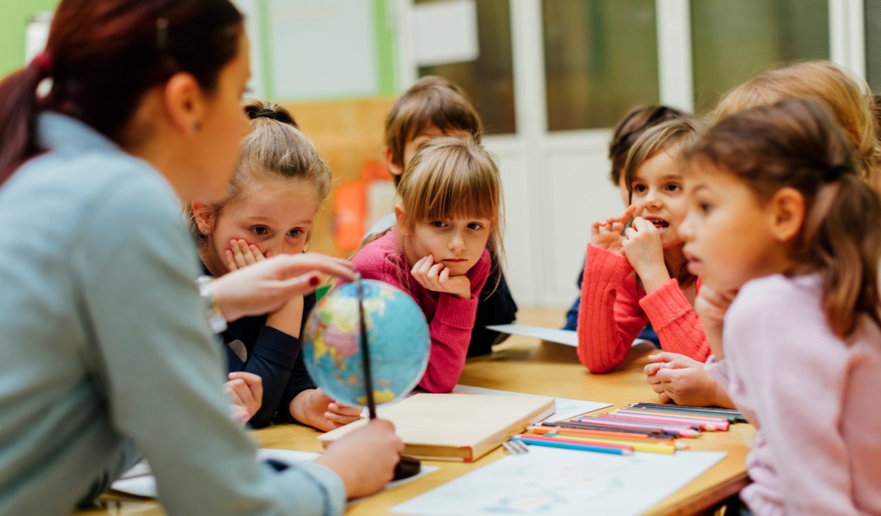 A female teacher sits at a table holding a small globe as she speaks to six captivated children who listed on.