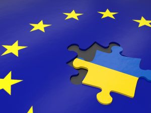 A puzzle piece marked with the blue and yellow Ukraine national flag sits out of place on a graphic of the European Union flag, a circle of yellow stars on a dark blue ground.