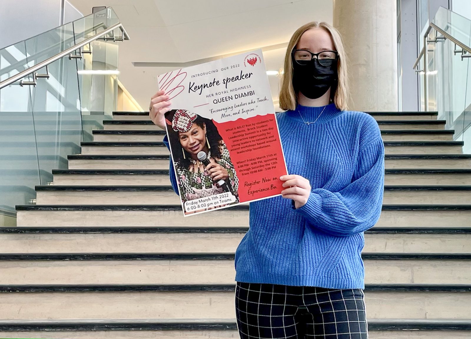 A masked woman in a blue sweater stands on a staircase while holding a poster that features information about Queen Diambi of the Democratic Republic of the Congo, who is pictured on the poster holding a microphone.