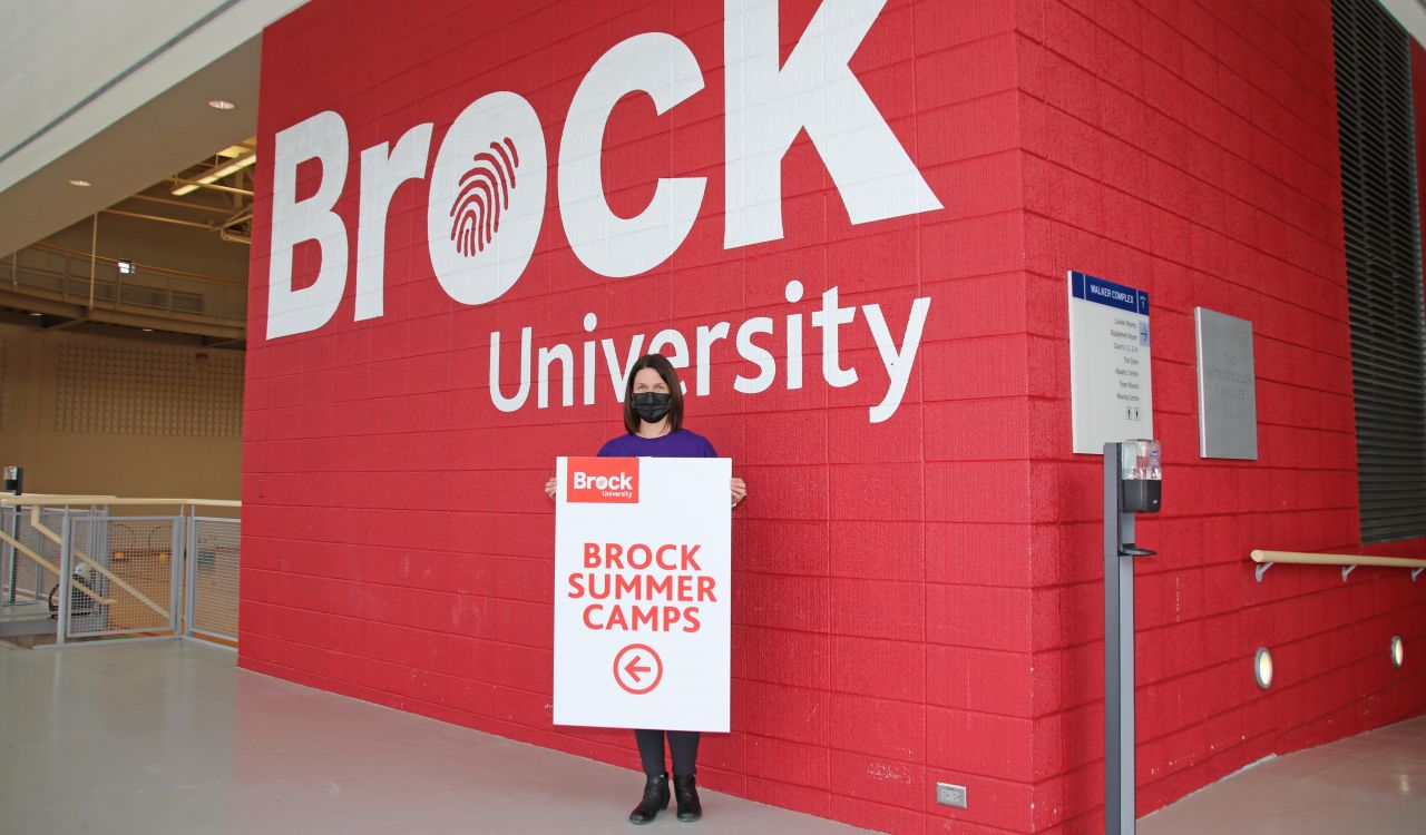 A masked woman in a purple shirt stands in front of a large red wall with ‘Brock University’ written on it in white letters. She is holding a sign with red letters on a white background that says ‘Brock Summer Camps.’