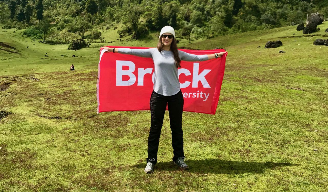 A young woman holds a red flag that reads 'Brock' while standing in the middle of a green field with trees lining the background.