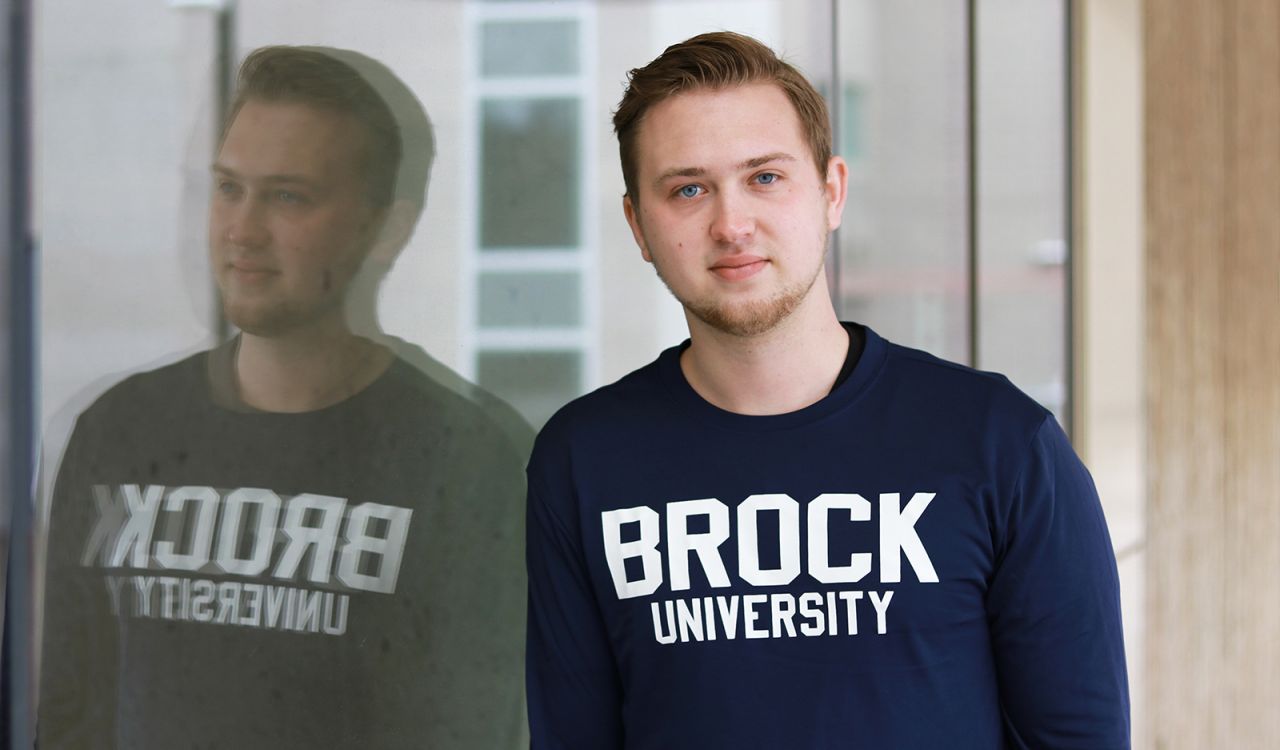 A young man leans against a glass wall, creating a reflection in the glass. He is wearing a blue long-sleeved shirt with ‘Brock University’ in white letters.