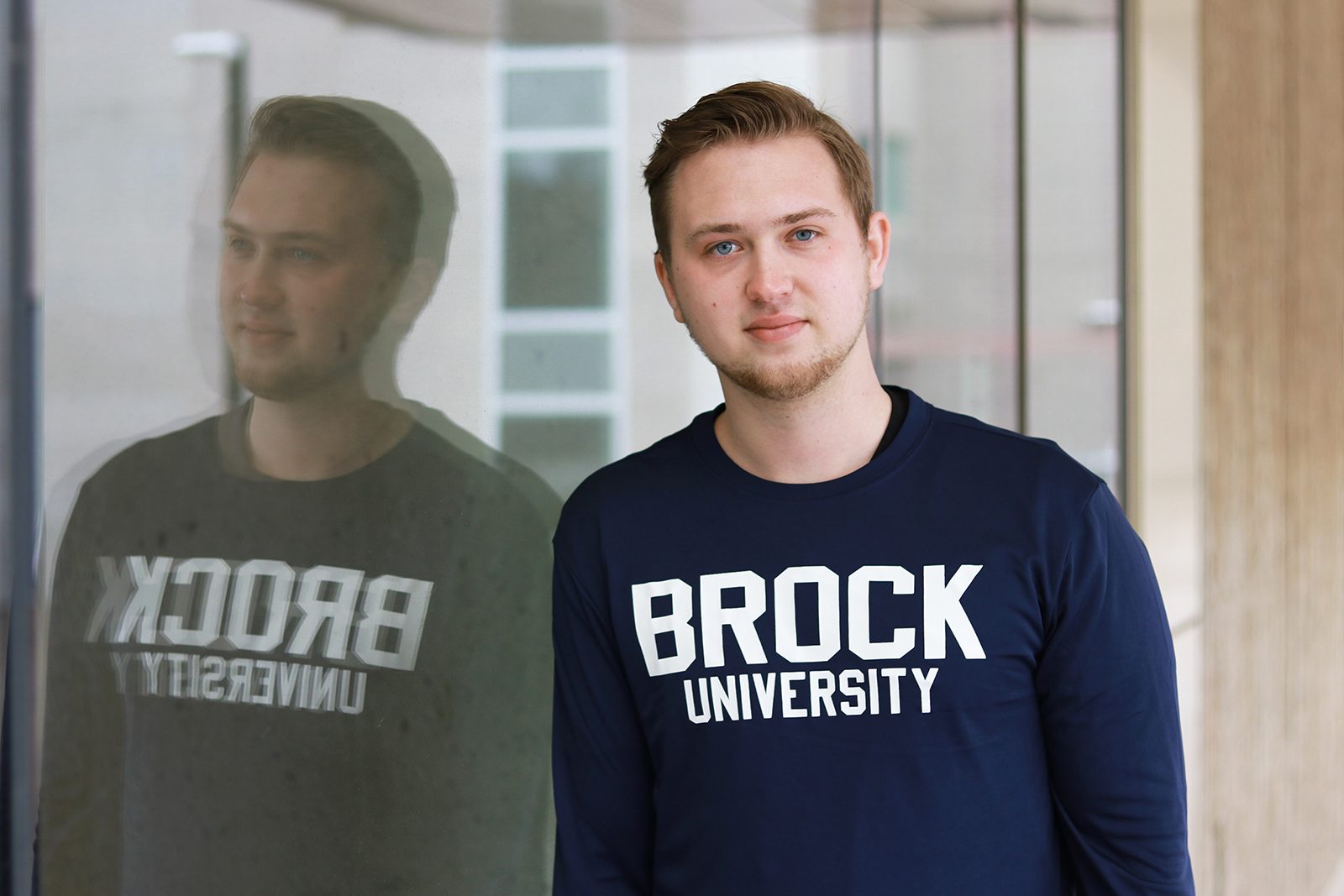 A young man leans against a glass wall, creating a reflection in the glass. He is wearing a blue long-sleeved shirt with ‘Brock University’ in white letters.