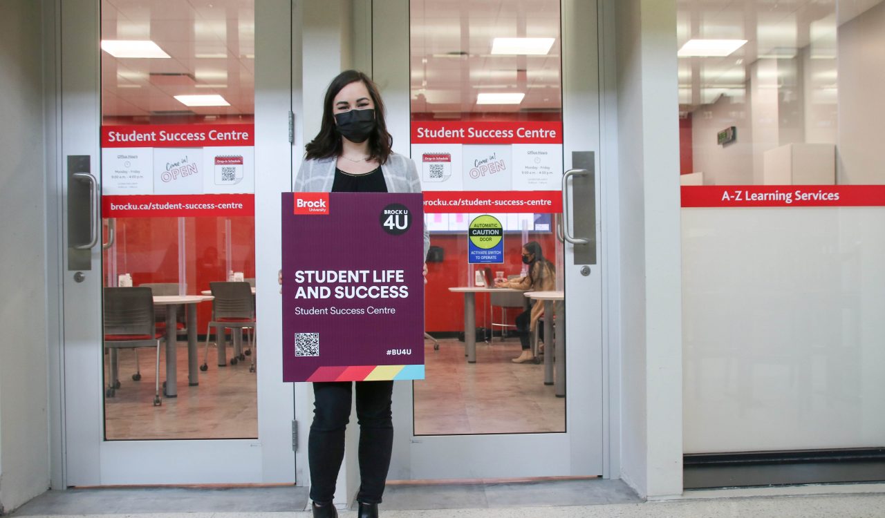 A woman holds a purple sign that says ‘Student Life and Success' in front of two glass doors.
