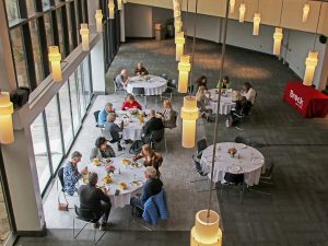Seventeen people eat lunch at round tables in a banquet room. Golden cylindrical lights hang from a high ceiling. A rectangular table covered in a red tablecloth with “Brock University” written on it can be seen near the tables.