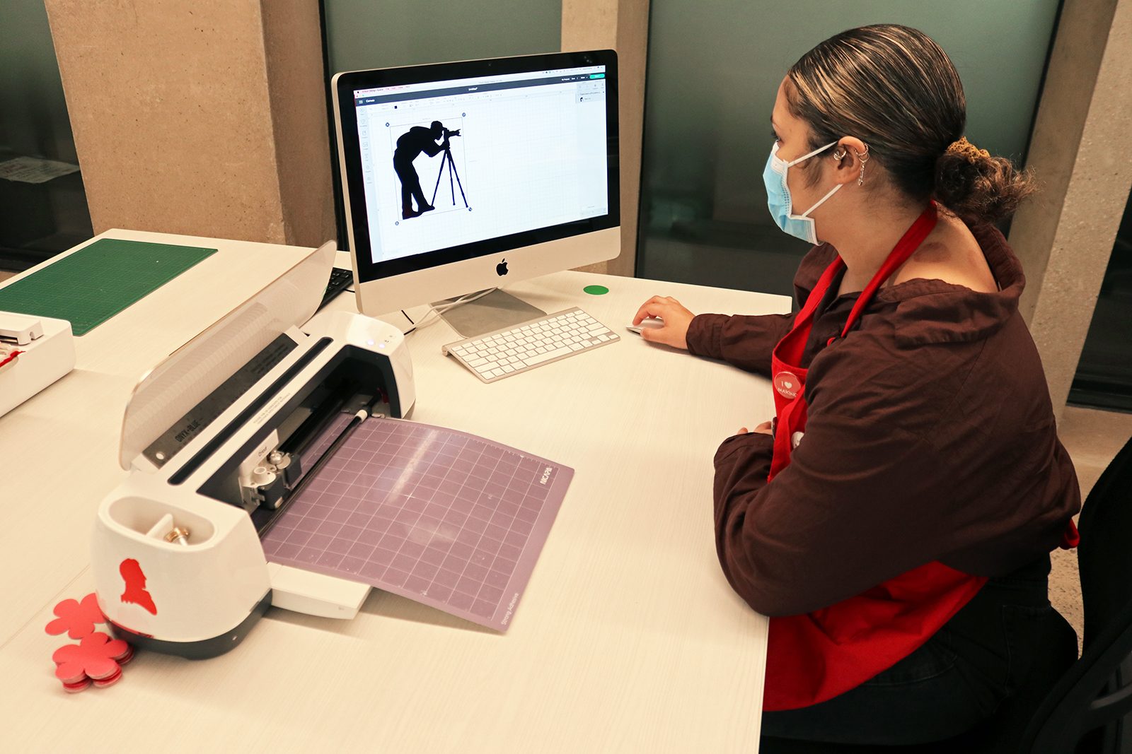 A woman wearing a red apron sits at a white table with her hand on a computer mouse. She looks at a large computer screen with a silhouetted illustration of a person bending over to use a camera on a tripod. Next to the desktop computer is a Cricut cutting machine.