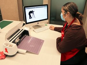A woman wearing a red apron sits at a white table with her hand on a computer mouse. She looks at a large computer screen with a silhouetted illustration of a person bending over to use a camera on a tripod. Next to the desktop computer is a Cricut cutting machine.