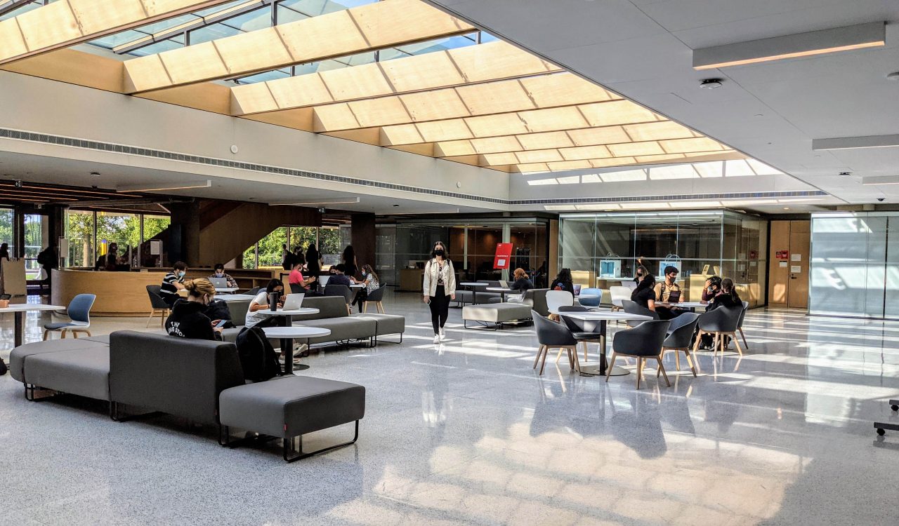 An indoor space, lit by a large skylight above, with people sitting at tables and walking by.