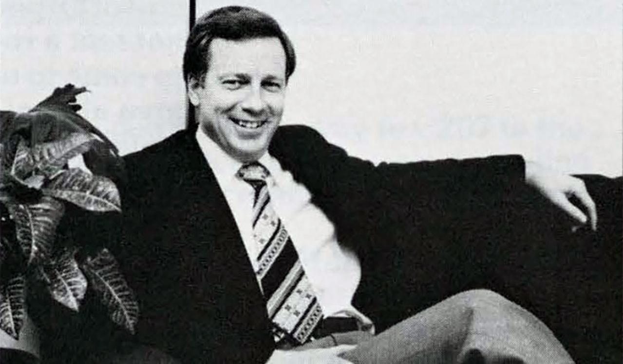 A black and white photo taken circa 1978 of a man sitting on a couch. The man is wearing a wearing a suit and tie. He is smiling, with one hard resting on the back of the couch and one hand on his crossed legs. A plant is to the left of him on the couch.