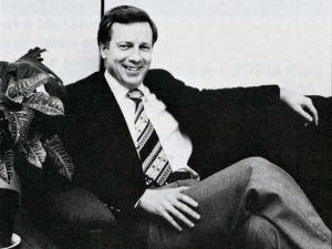 A black and white photo taken circa 1978 of a man sitting on a couch. The man is wearing a wearing a suit and tie. He is smiling, with one hard resting on the back of the couch and one hand on his crossed legs. A plant is to the left of him on the couch.
