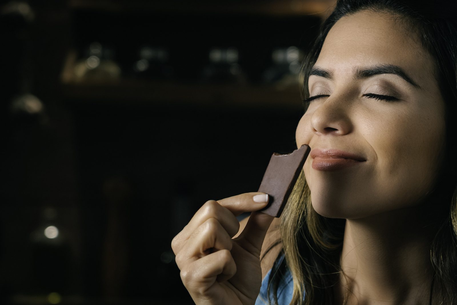 A woman holds up a piece of dark brown chocolate to her nose. Her eyes are closed and she has a smile on her face as she smells it.