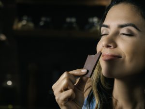 A woman holds up a piece of dark brown chocolate to her nose. Her eyes are closed and she has a smile on her face as she smells it.