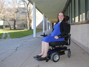 A woman in a purple dress sits in a motorized wheelchair under a covered outdoor walkway.