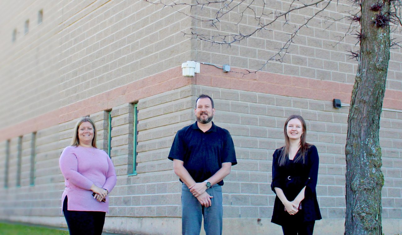 Amanda Smits, ESRC Centre Administrator, Dave O’Connor, BUSU Director of Food and Beverage, and Kayleigh Rossetto, BUSU Director of Governance and Advocacy, stand on the grass next to a row of trees in front of the Brock Student-Alumni Centre.