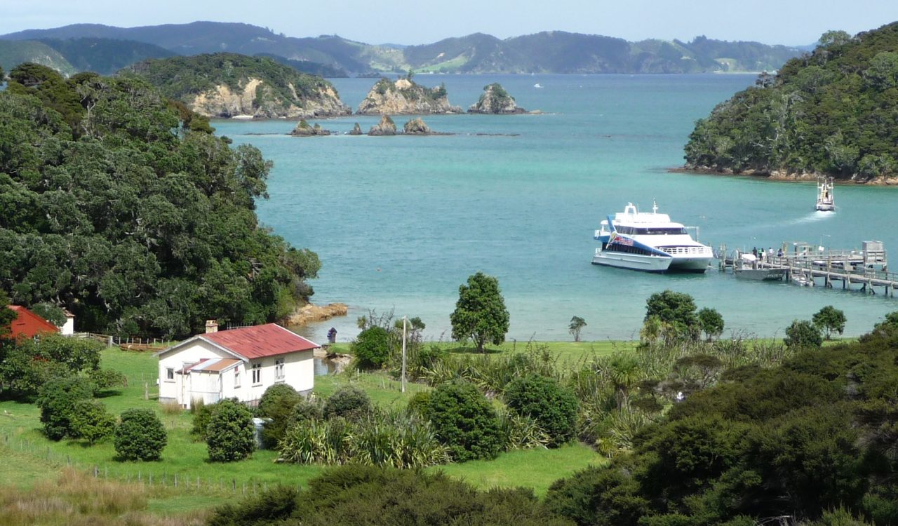 A scene of the Bay of Islands in New Zealand showing a white house with a red roof on the grass next to the water, where a white boat is moored at a dock and people wait to board it.