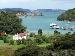 A scene of the Bay of Islands in New Zealand showing a white house with a red roof on the grass next to the water, where a white boat is moored at a dock and people wait to board it.