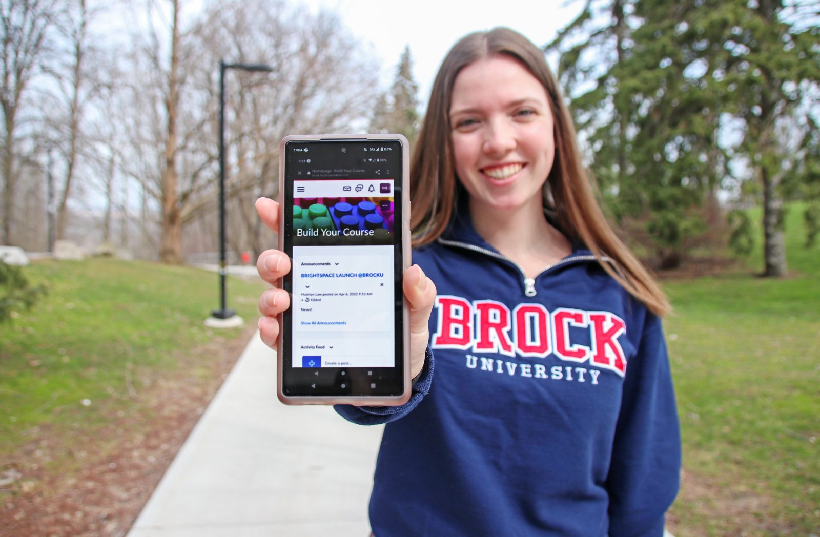 A young woman in a Brock University sweater holds up a smartphone with her arm outstretched and the screen facing the camera.