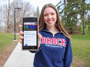 A young woman in a Brock University sweater holds up a smartphone with her arm outstretched and the screen facing the camera.