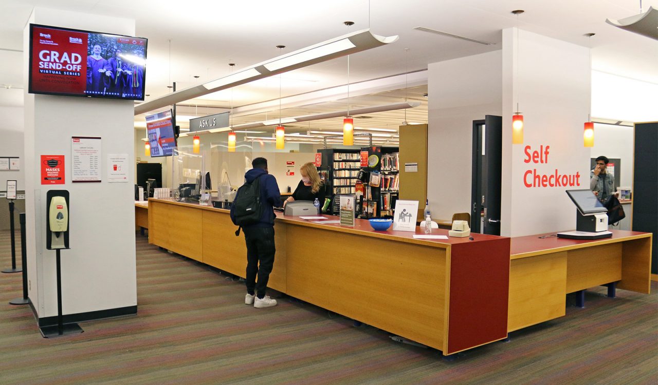 A student stands at a long desk facing another person who is helping him with library material. A sign hangs over the desk that reads ‘ask us.’ Behind the desk are two rows of tall shelves filled with books. A second, shorter desk offers self-checkout of library materials.