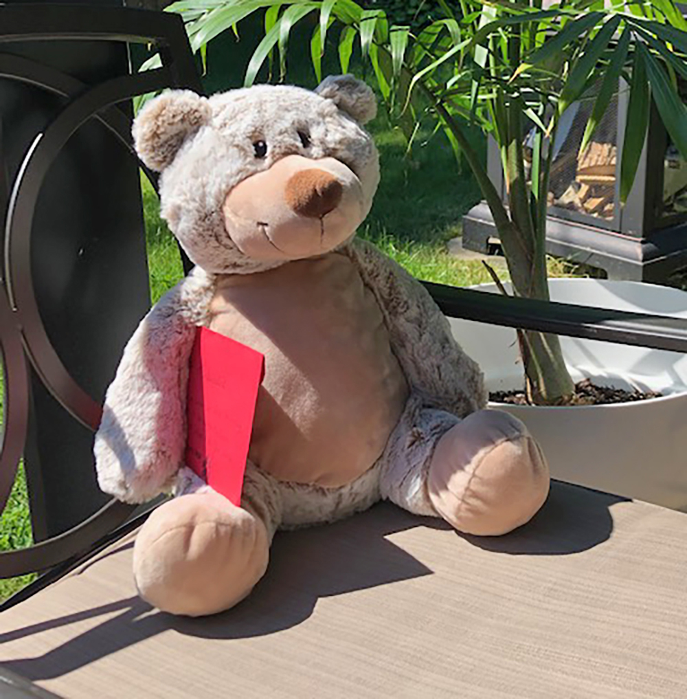 A brown teddy bear holding a red envelope is seated on a chair outside.