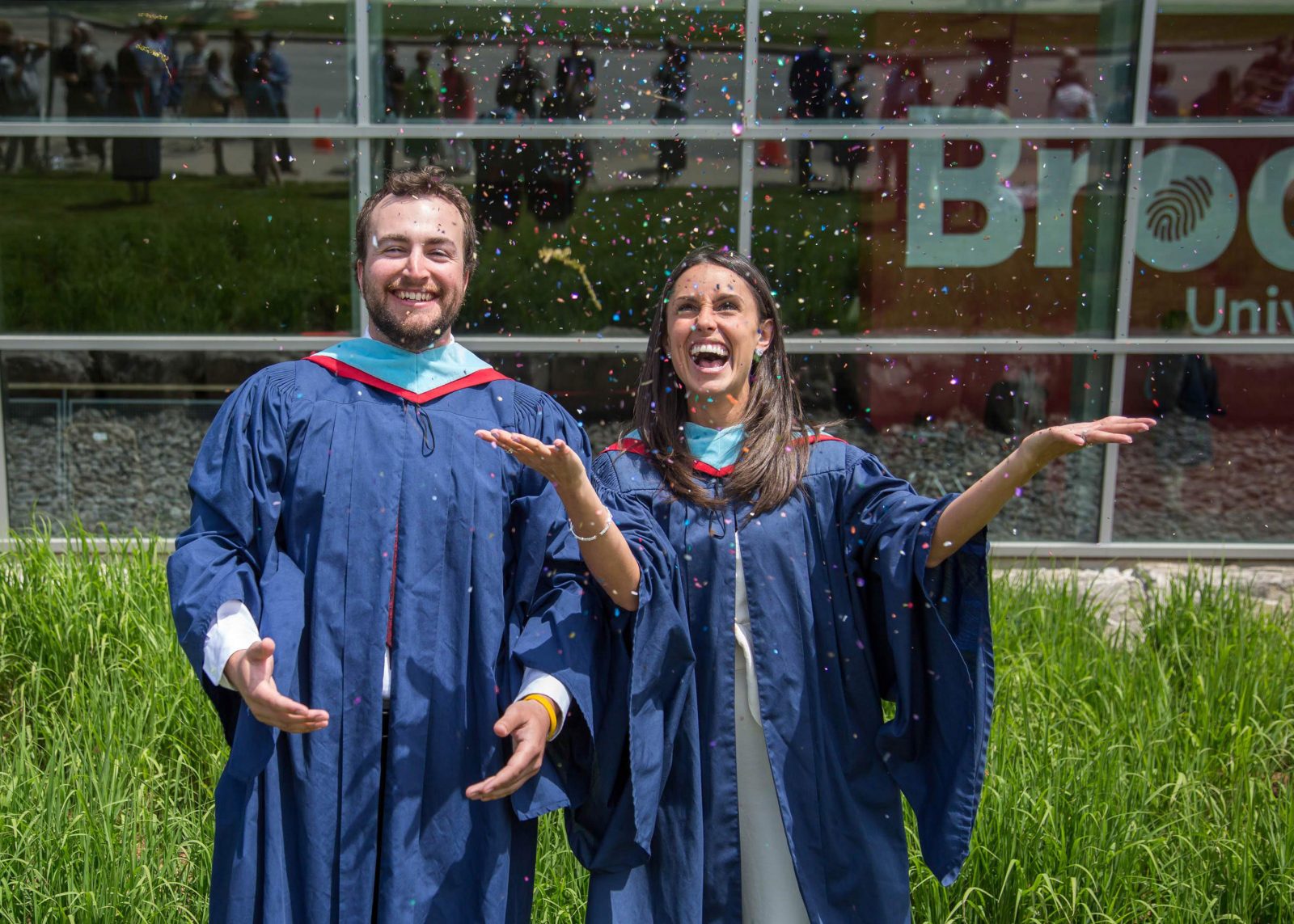 One male and one female university graduate throw confetti in the air while standing in front of a glass building.