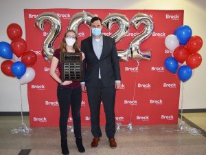 A woman and a man stand together in front of a red backdrop with white ‘Brock University’ repeated several times. Red, white and blue balloons flank them on either side. Silver ‘2022’ balloons float behind them. The woman on the left holds a large plaque. Both are wearing a mask.