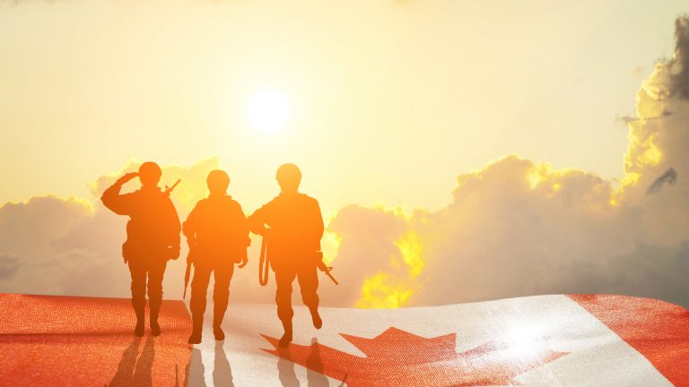 The silhouette of three soldiers standing on a field made up of a Canadian flag with the sunset in the background.