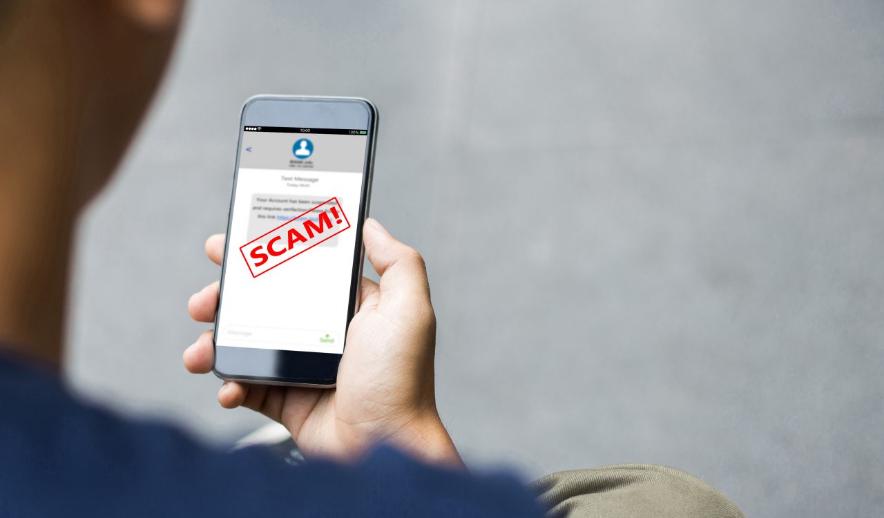 A man holds a smartphone with the word "Scam!" written across the screen in bold red letters.