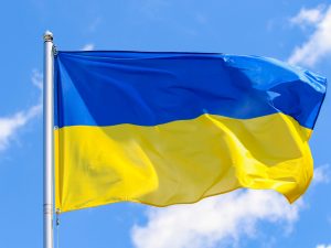 The Ukrainian flag, with blue on the top and yellow on the bottom, flies on the right side of a flagpole against a blue sky.