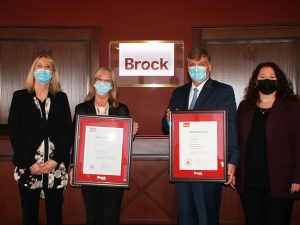 Four people stand next to each other in front of a glass sign that reads “Brock.” The two people in the middle hold framed certificates.