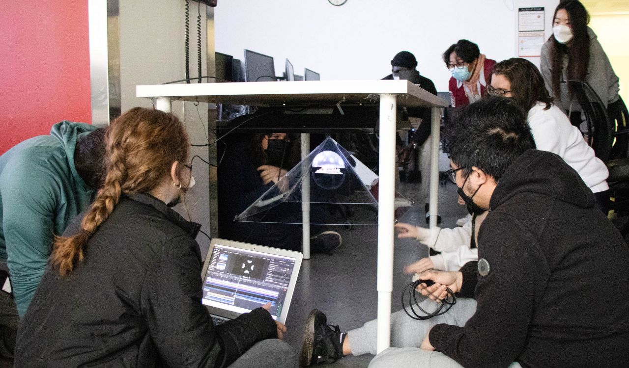 Students sitting on the floor around a table that has a plexiglass pyramid suspended underneath. The room is darkened and an animal can be seen floating in the pyramid.