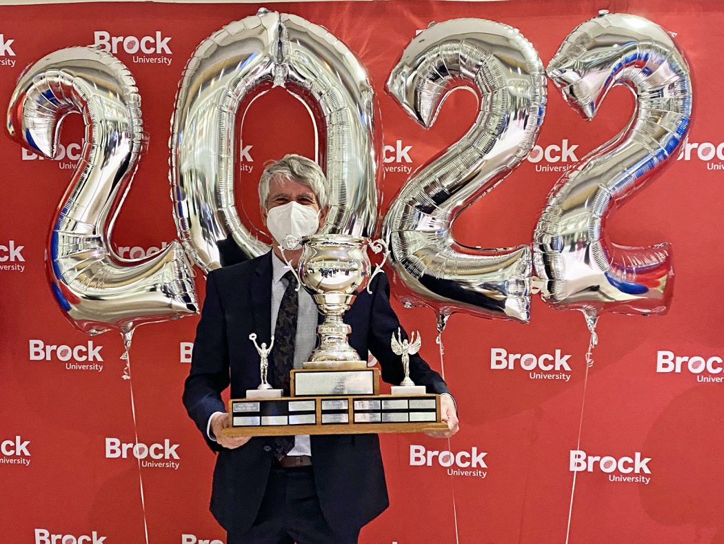 A man holding a large trophy stands in front of a red backdrop with white ‘Brock University’ repeated several times. Silver ‘2022’ balloons float behind him.