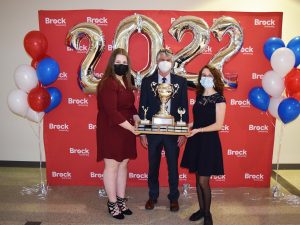 Three people stand together in front of a red backdrop with white ‘Brock University’ repeated several times. The person in the centre holds a large trophy. Silver ‘2022’ balloons float behind them. Everyone is wearing a mask.