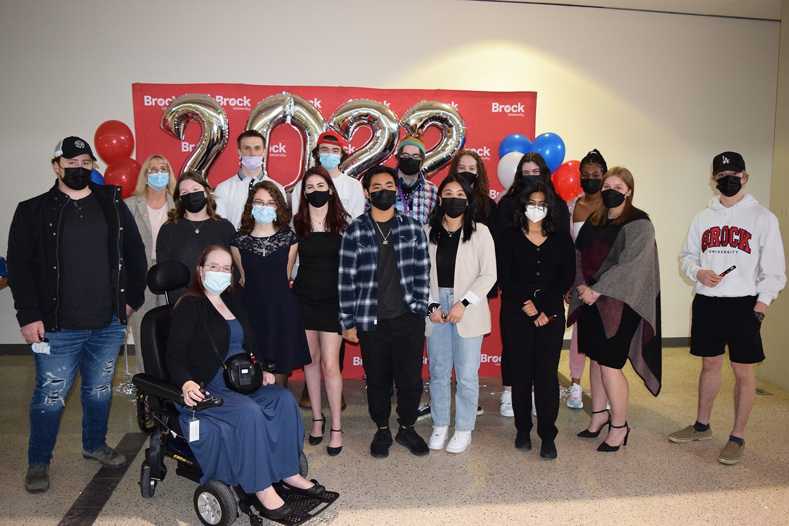 Seventeen people stand together in two rows in front of a red backdrop with white ‘Brock University’ repeated several times. Silver ‘2022’ balloons float behind them. Everyone is wearing a mask.