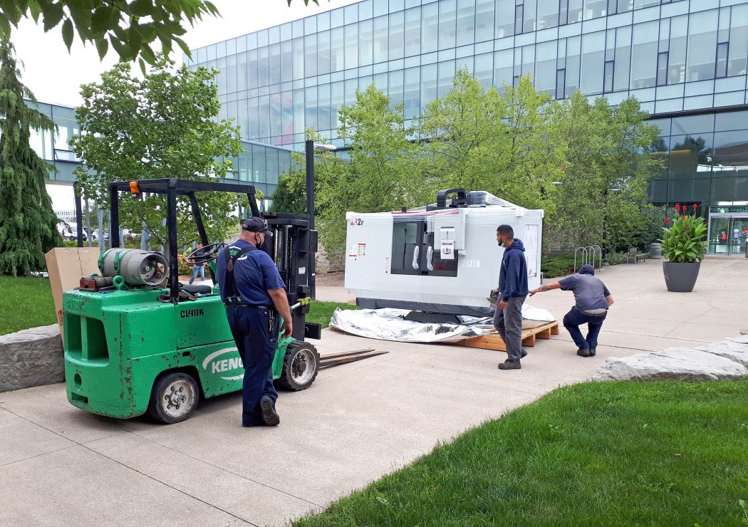 Three men unload a waterjet machine using a forklift and skid outside on a concrete concourse. A glass building with trees and green foliage appear in the background.