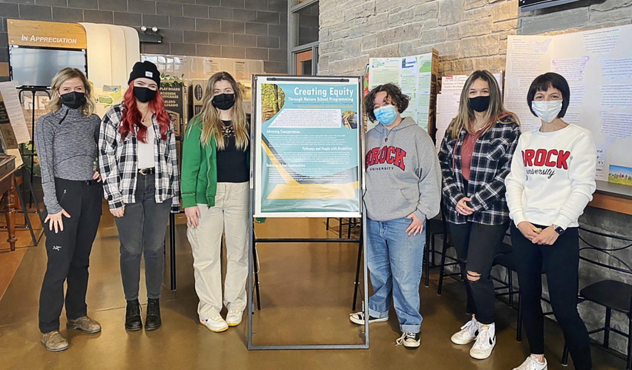 Six masked people stand next to a student-made poster displayed in a large room.