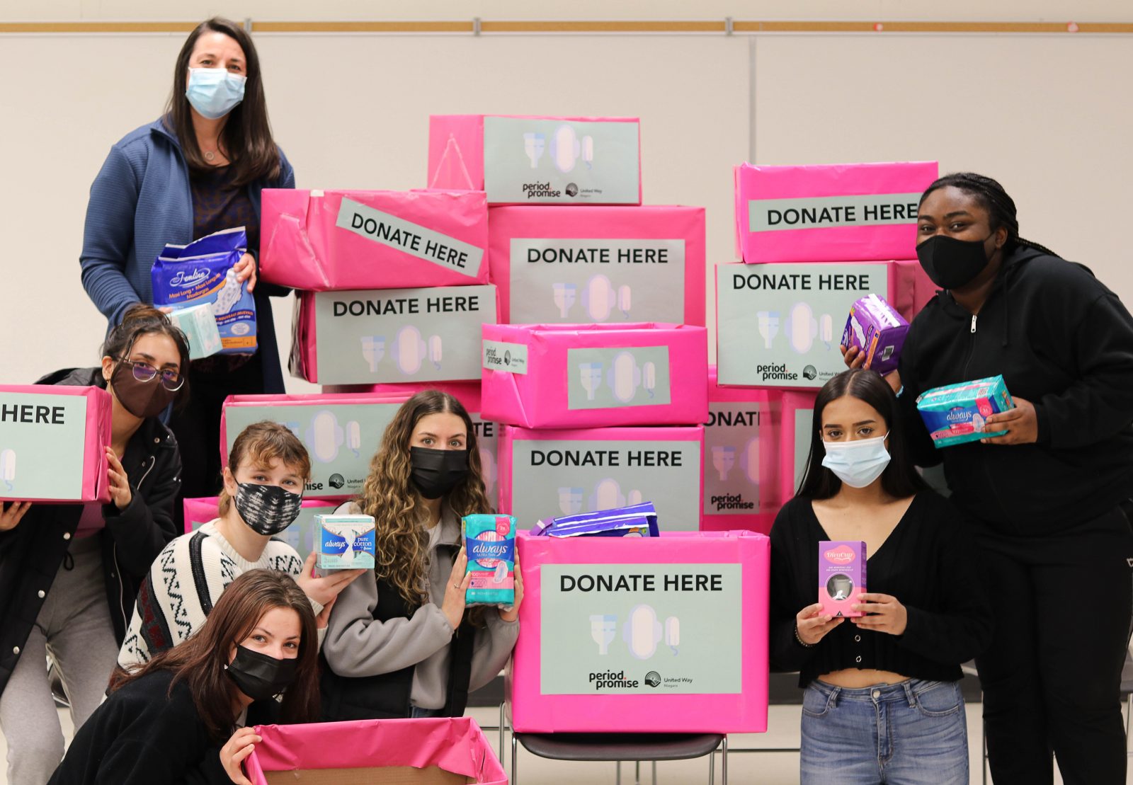 Seven masked women stand around pink boxes with signs that say "Donate here" on them.