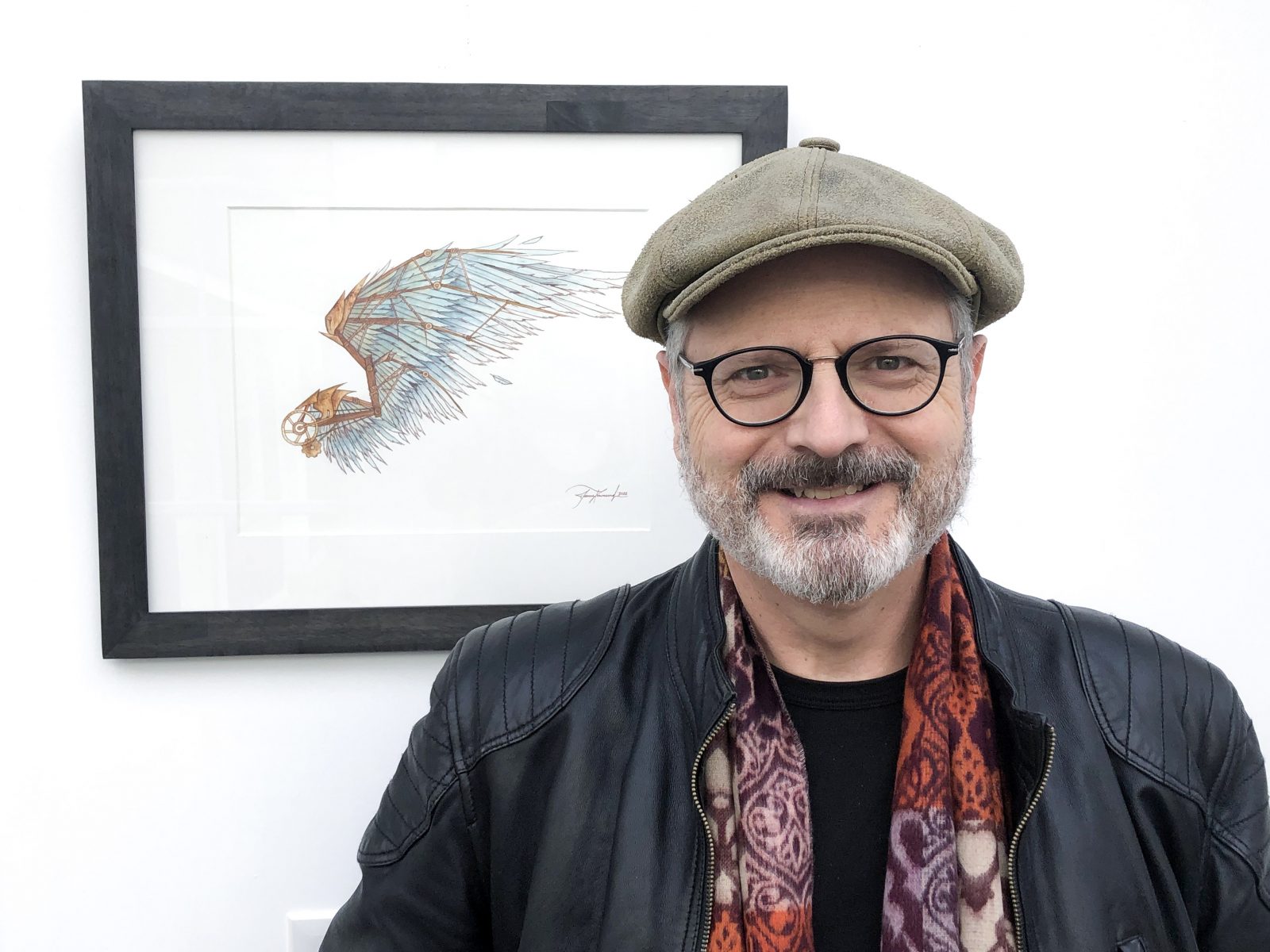 A portrait of Phil Mackintosh in a leather jacket and hat in front of artwork depicting a manufactured wing.