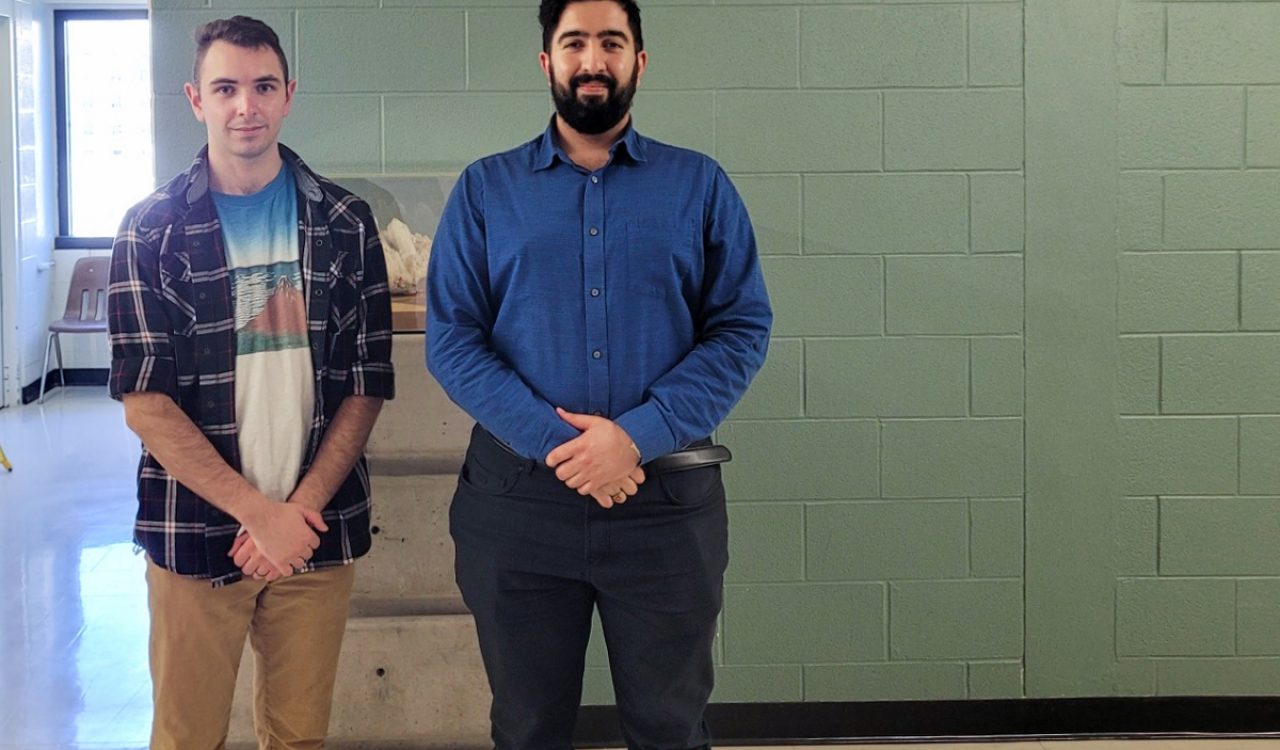 Two Brock University students pose in front of the Earth Sciences department sign. Pierre Simiganoschi stands to the left wearing plaid shirt with graphic t-shirt underneath and khaki pants. Nima Vaez-zadeh Asadi stands on the right wearing a blue dress shirt and dark dress pants.