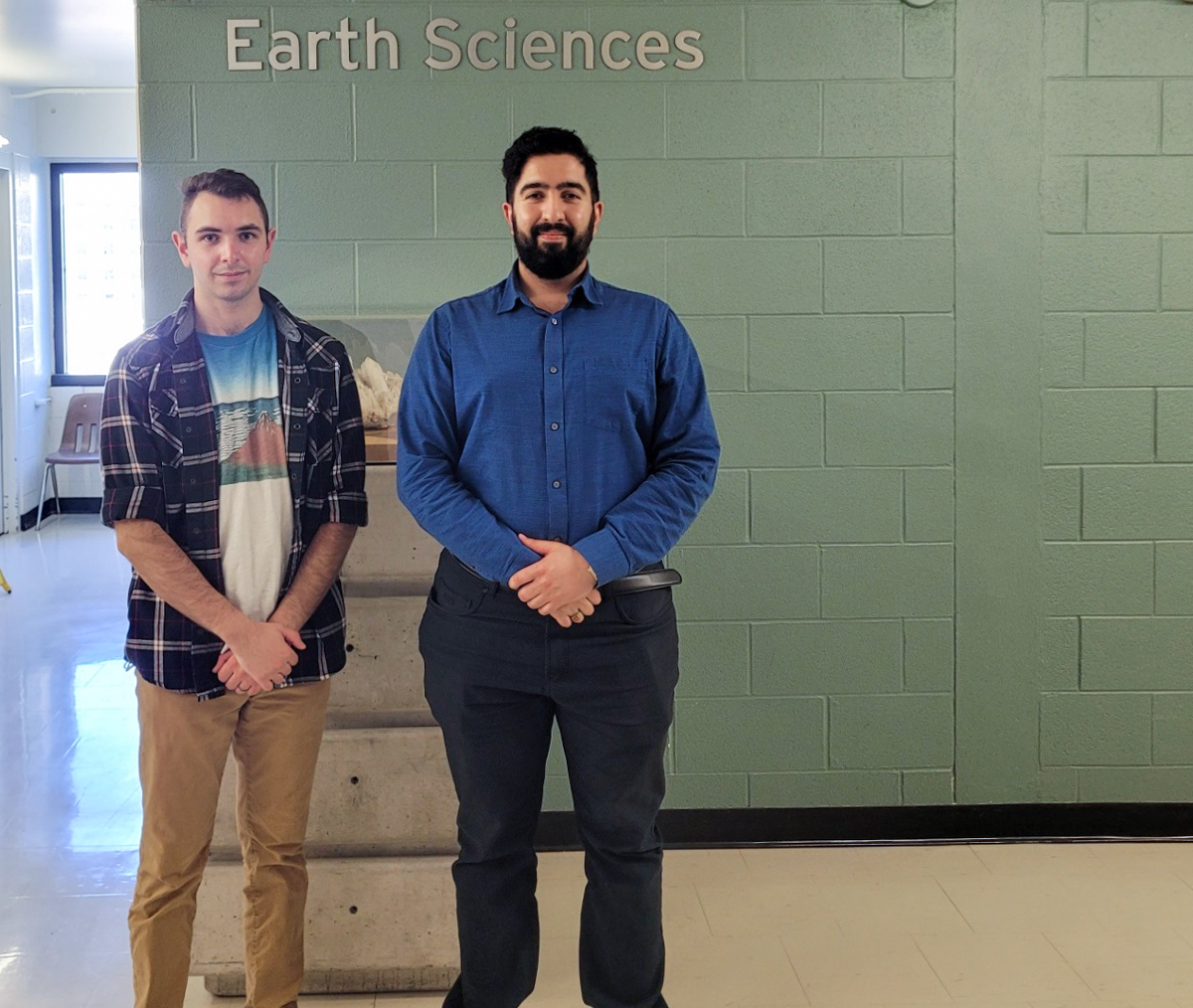 Two Brock University students pose in front of the Earth Sciences department sign. Pierre Simiganoschi stands to the left wearing plaid shirt with graphic t-shirt underneath and khaki pants. Nima Vaez-zadeh Asadi stands on the right wearing a blue dress shirt and dark dress pants.