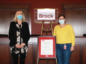 Two woman stand near each other. In between them is a gold easel with a framed certificate.